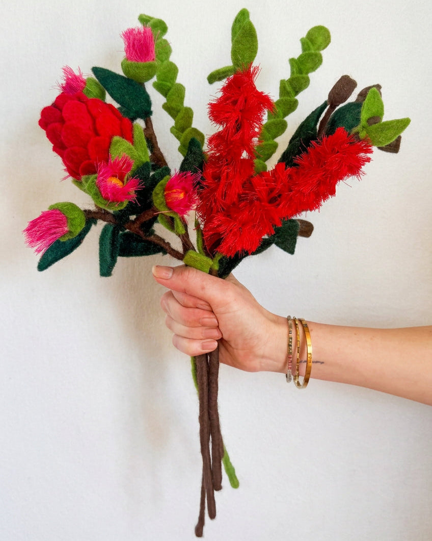 A hand holding a handmade felt flower bouquet with bright red and pink blossoms, green leaves, and brown stems against a plain white wall.