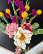 A hand holds a colorful felt flower bouquet with a large white daisy, pink rose, white and pink flowers, yellow flower buds, and green stems against a dark background.
