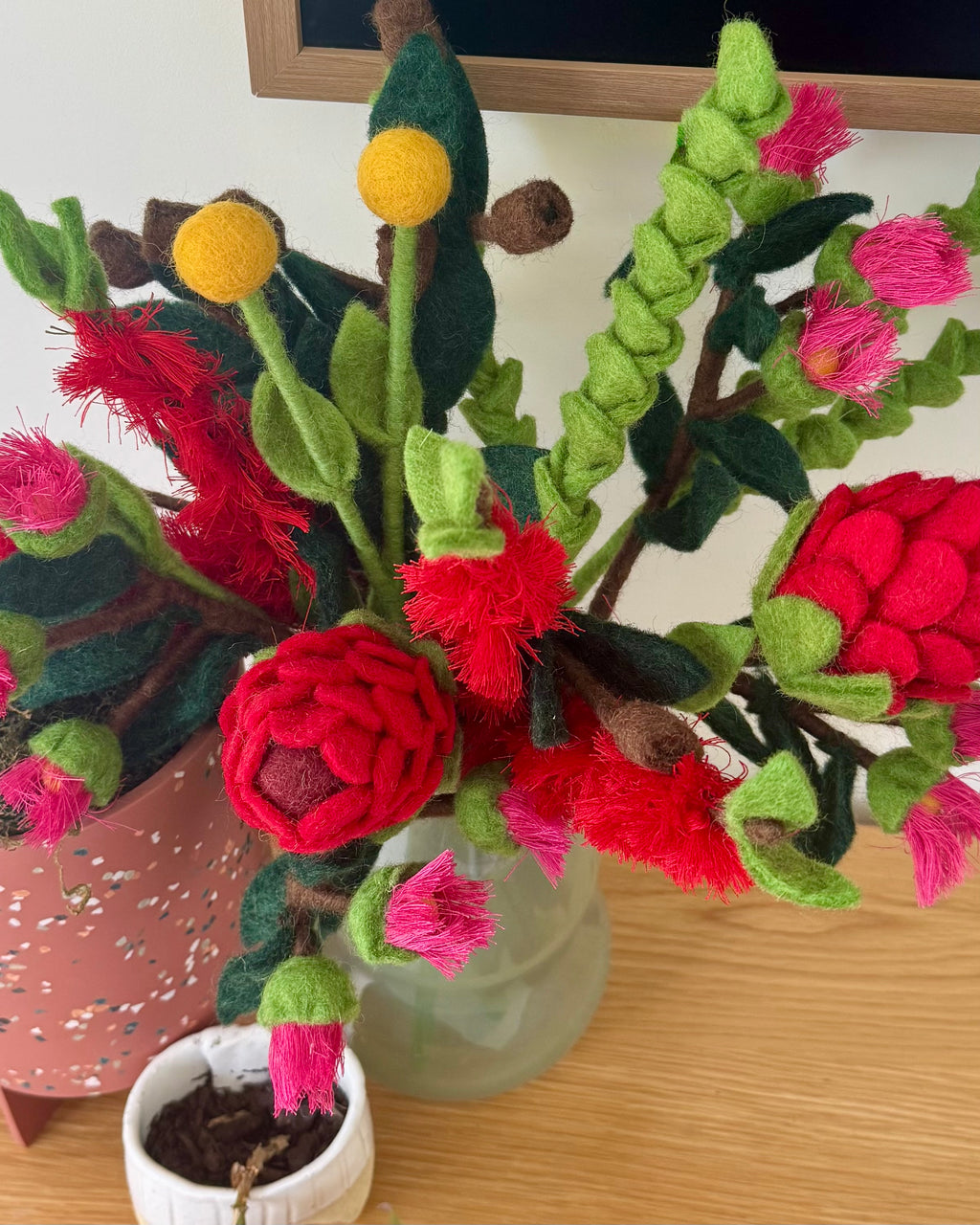 A handmade felt flower arrangement in a frosted glass vase, featuring bright red waratah blooms, pink buds, green leaves, and yellow flower tops on a wooden tabletop. All australian flowers
