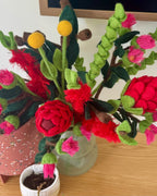 A handmade felt flower arrangement in a frosted glass vase, featuring bright red waratah blooms, pink buds, green leaves, and yellow flower tops on a wooden tabletop. All australian flowers
