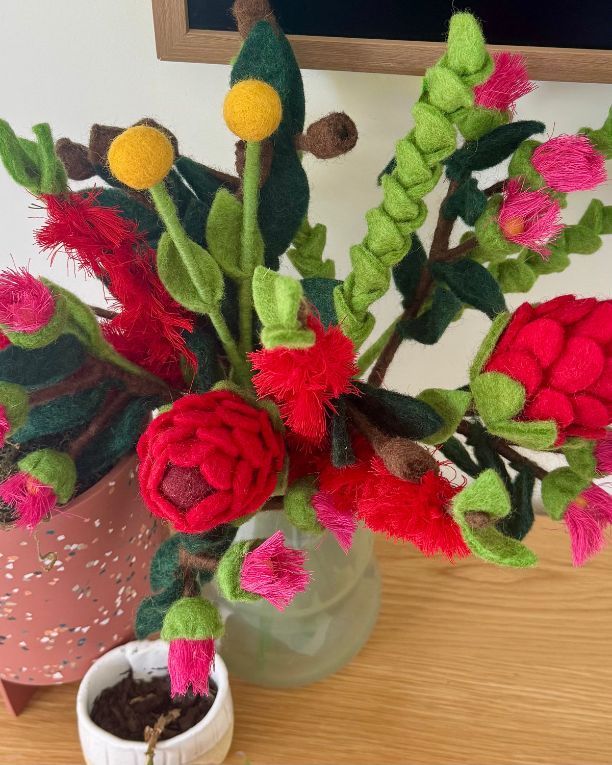 A handmade felt flower arrangement in a frosted glass vase, featuring bright red waratah blooms, pink buds, green leaves, and yellow flower tops on a wooden tabletop. All australian flowers
