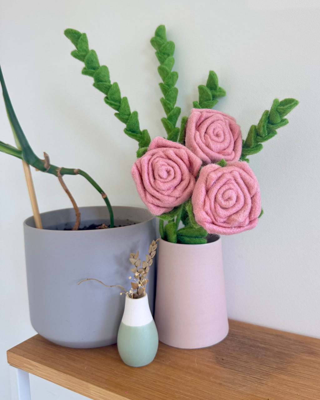 Three pink felt roses with green leaves arranged in a light pink cylindrical vase on a wooden shelf, next to a gray planter and a small mint green vase.