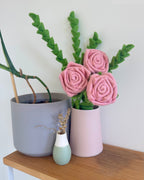 Three pink felt roses with green leaves arranged in a light pink cylindrical vase on a wooden shelf, next to a gray planter and a small mint green vase.