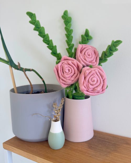 Three pink felt roses with green leaves arranged in a light pink cylindrical vase on a wooden shelf, next to a gray planter and a small mint green vase.