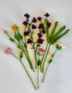 A set of handmade felt flower stems arranged on a white fabric background, featuring dark purple sweet pea, yellow billy buttons, pink rose and lily, and white daisy with green leaves and stems.