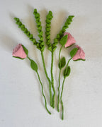 A set of small artificial flower stems laid flat on a white textured background, including pink fabric rose buds and green leafy sprigs and stems.