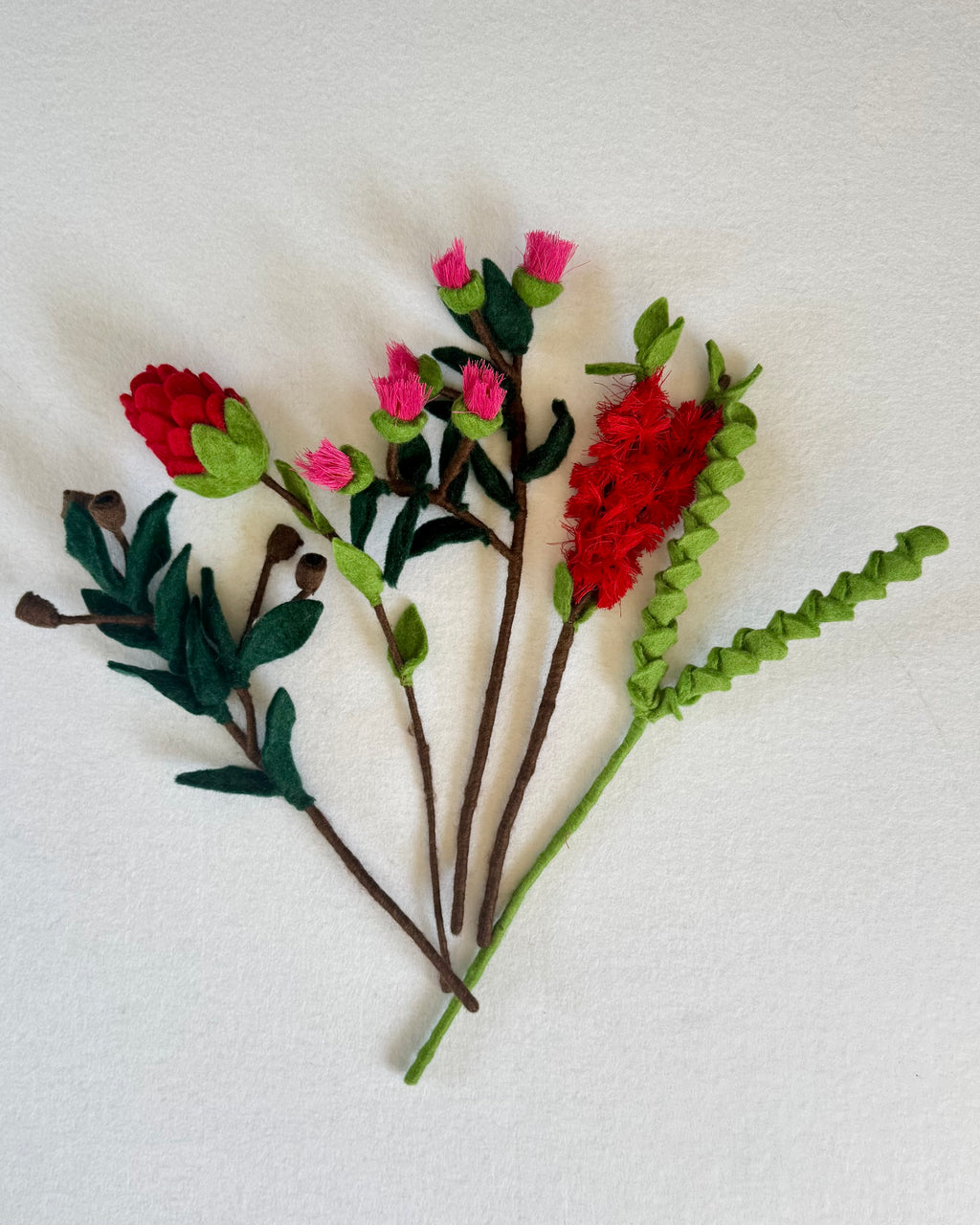 A set of handmade felt artificial flowers and leaves on brown stems, featuring red and pink blossoms with green leaves, laid out on a white textured fabric background. It consits of Australian flora.