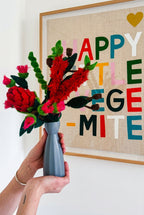 A hand holds a gray ceramic vase filled with a colorful handmade australian felt flower arrangement, set against a white wall with a framed 'HAPPY LITTLE VEGE MITE' artwork.