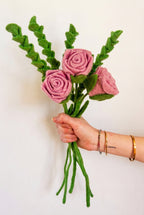 A hand holding a small felt bouquet of pink roses with green leaves and stems against a plain light wall.