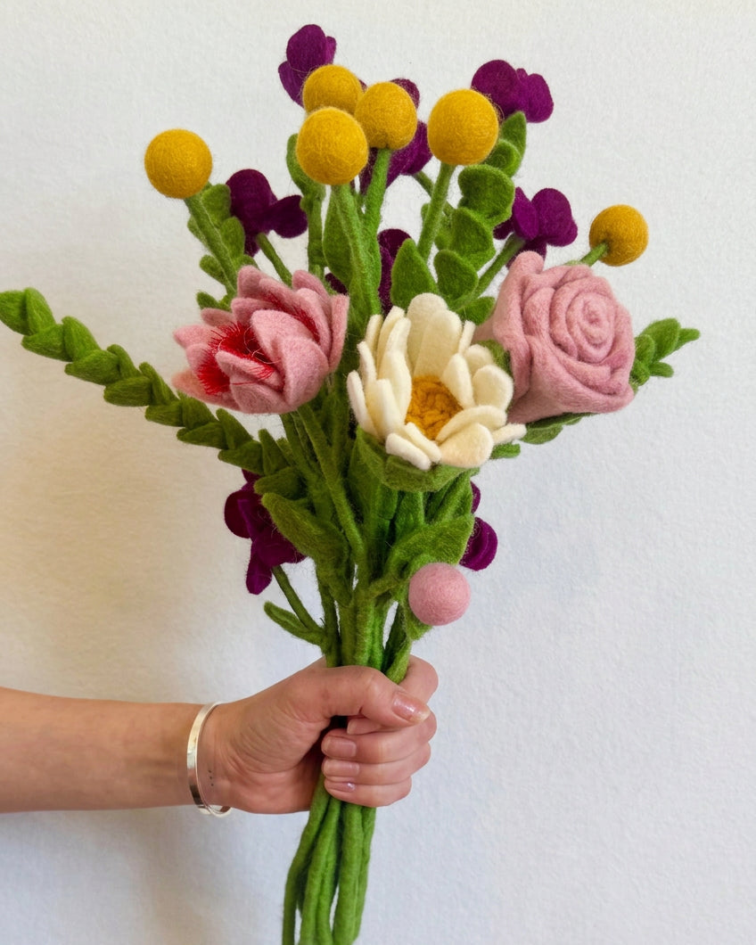 A hand holding a handmade felt flower bouquet with pink roses, a white daisy, yellow pom-pom balls, and purple blossoms against a plain white wall.