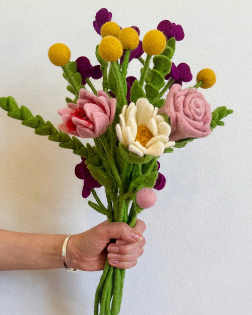 A hand holding a handmade felt flower bouquet with pink roses, a white daisy, yellow pom-pom balls, and purple blossoms against a plain white wall.