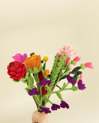 A hand holding a small felt flower bouquet with red, orange, pink, purple, and green felted wool flowers against a plain beige background.