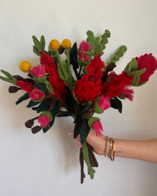 A hand holding a handmade felt flower bouquet with red and pink flowers, green leaves, and small yellow accents against a plain white wall.