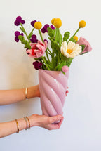 A person's hands holding a glossy, twisted light pink ceramic vase filled with a colorful felt flower bouquet against a plain white wall.