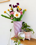 A pink spiral-patterned glass vase filled with a colorful handmade felt flower arrangement, styled on a wooden shelf next to potted green plants against a white wall.