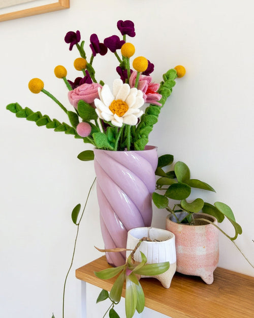 A pink spiral-patterned glass vase filled with a colorful handmade felt flower arrangement, styled on a wooden shelf next to potted green plants against a white wall.