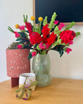 A colorful handcrafted felt floral arrangement in a pale green glass vase, displayed on a wooden table alongside a terrazzo planter and a small potted air plant. Australian native flowers and foilage.