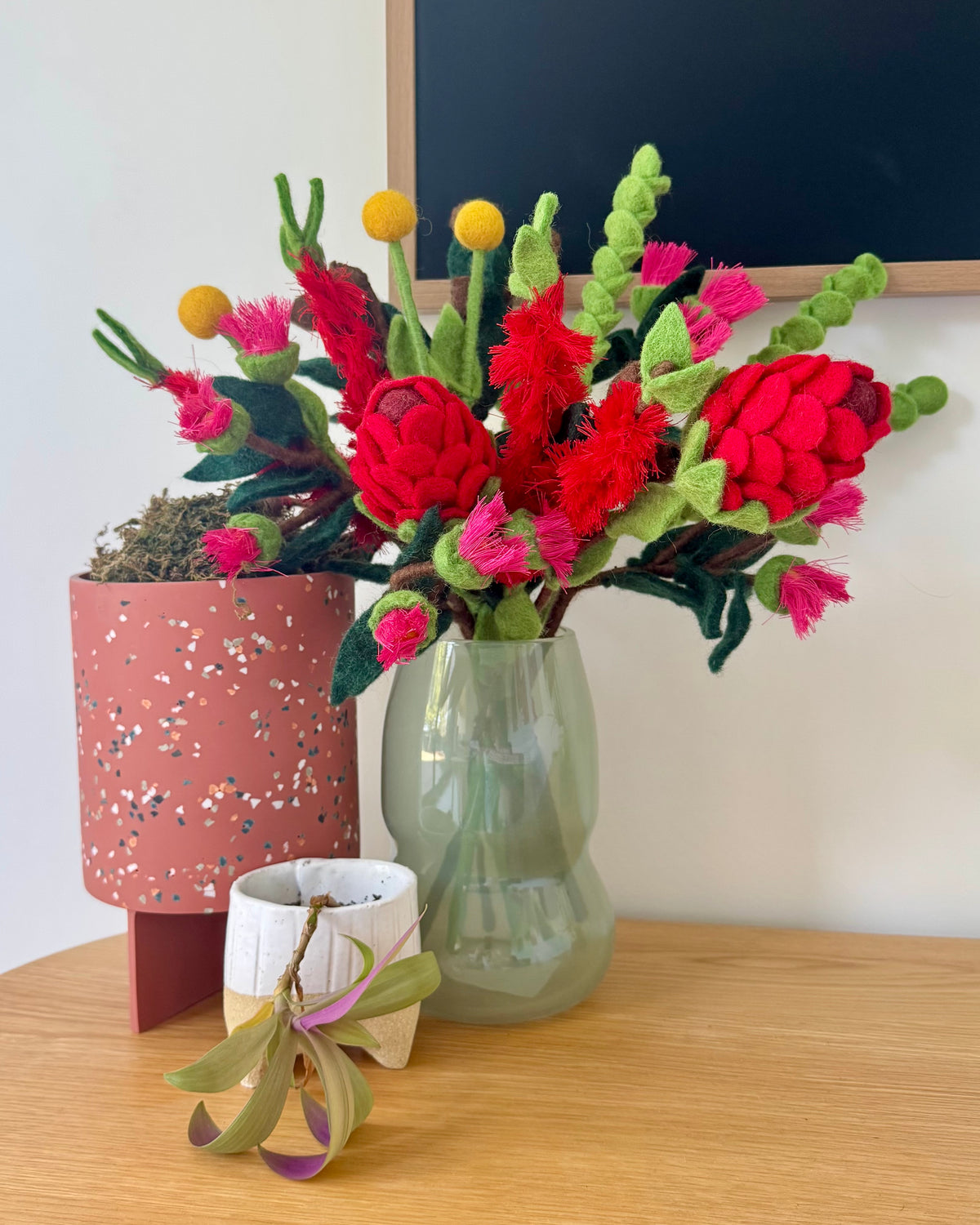 A colorful handcrafted felt floral arrangement in a pale green glass vase, displayed on a wooden table alongside a terrazzo planter and a small potted air plant. Australian native flowers and foilage.