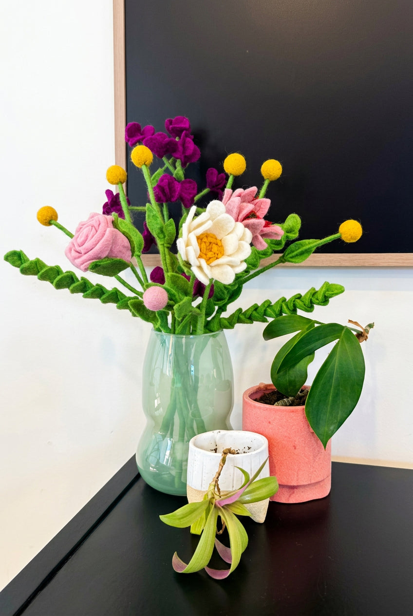 A handmade felt flower arrangement in a light green glass vase, displayed on a dark table next to a potted green plant. The felt bouquet features pink roses, a white daisy with yellow center, purple blossoms, and yellow globe flowers.