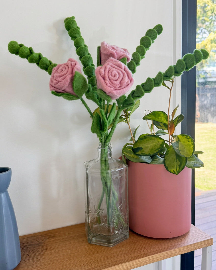 A decorative arrangement of pink felt roses with green felt stems and leaves, displayed in a clear glass bottle vase next to a pink planter with a green houseplant, set on a wooden table near a window.