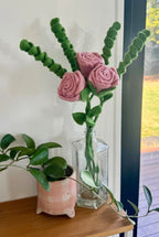 A handmade felt flower bouquet with three pink felt roses and green felt fern stems, arranged in a clear glass vase on a wooden shelf next to a potted plant.