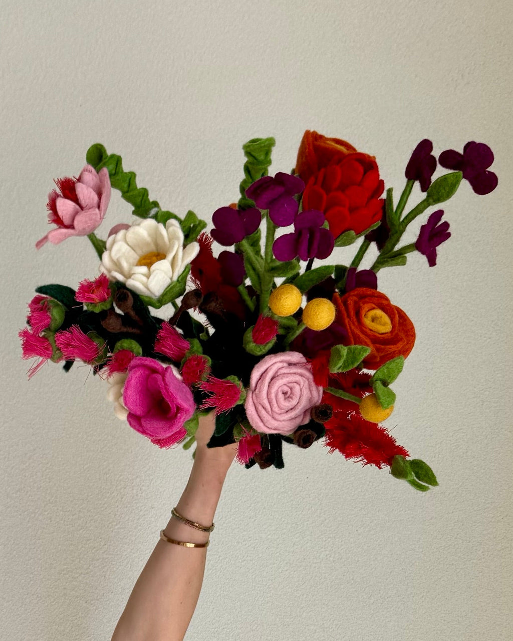 A hand holding a vibrant, multicolor artificial flower bouquet against a plain beige wall. The bouquet features a mix of red, pink, purple, white, and yellow flowers with green leaves.