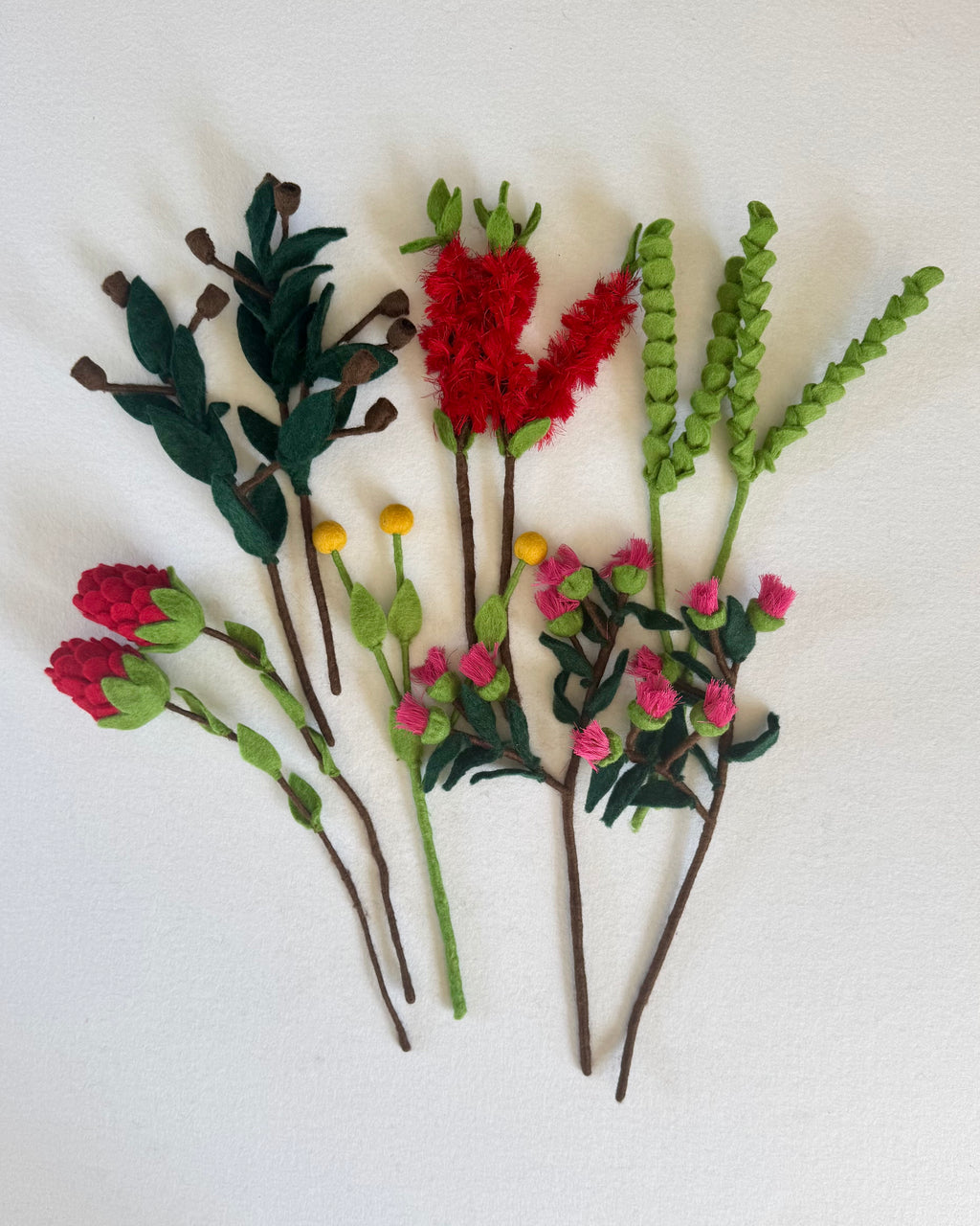 A set of handmade Australian felt floral stems arranged on a white background, featuring red and pink flowers, green leaves, and brown stems.