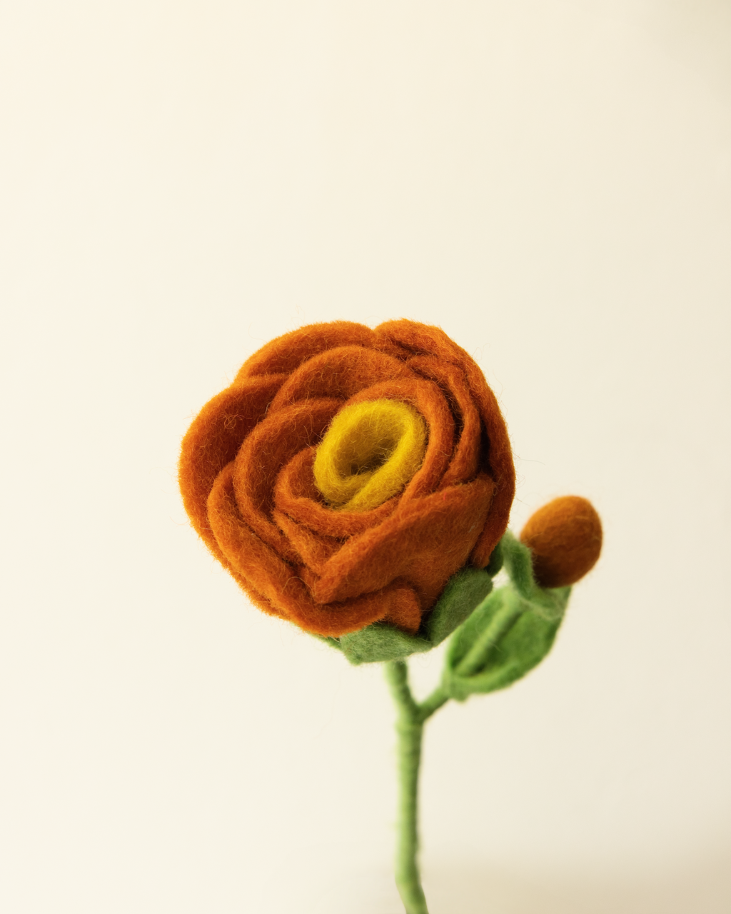 A hand holds a felted orange rose stem with a yellow center, green leaves, and a small bud against a plain off-white background.