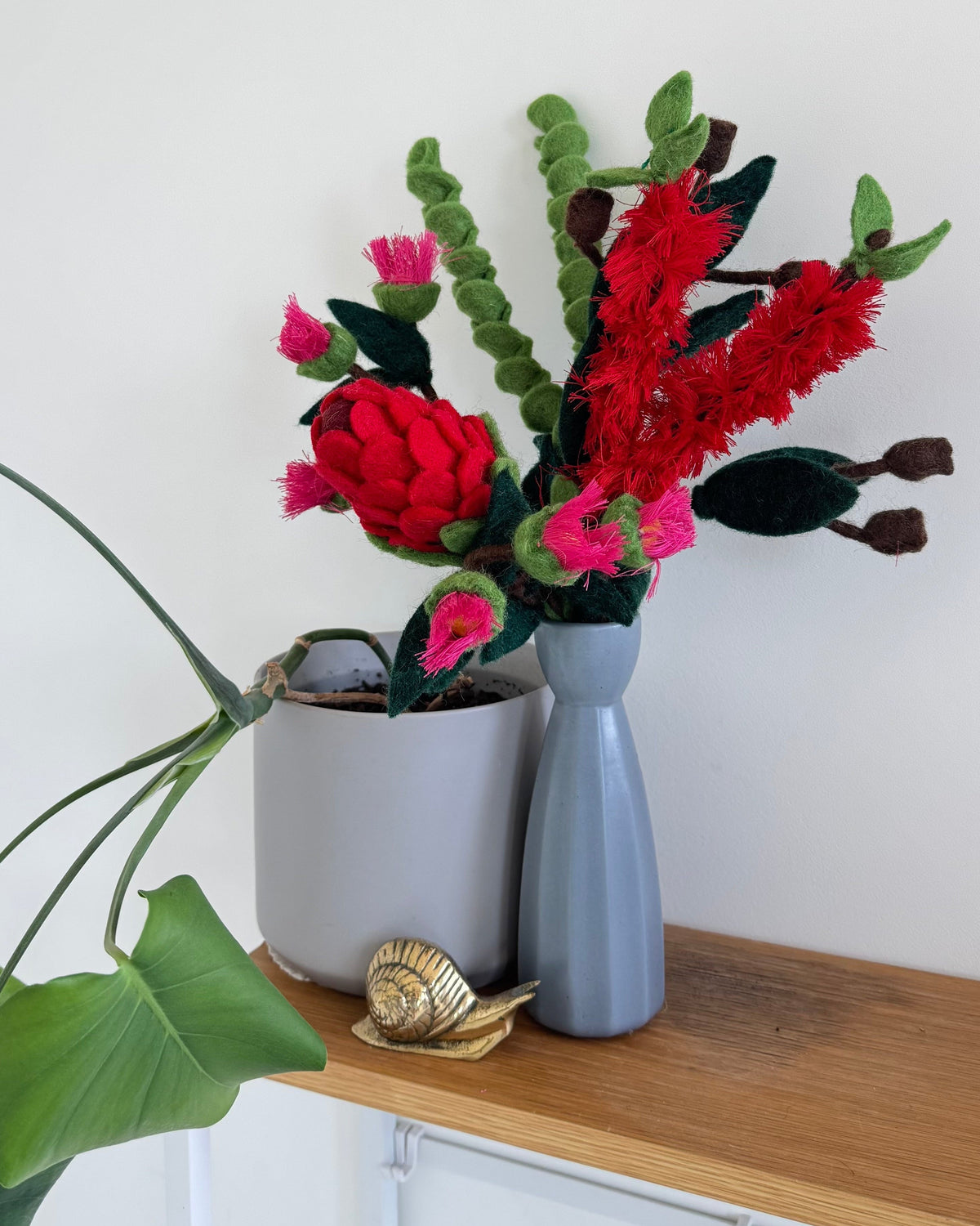 A styled arrangement of australian themed felt red and pink flowers with green leaves in a vase, placed on a wooden console table against a white wall.