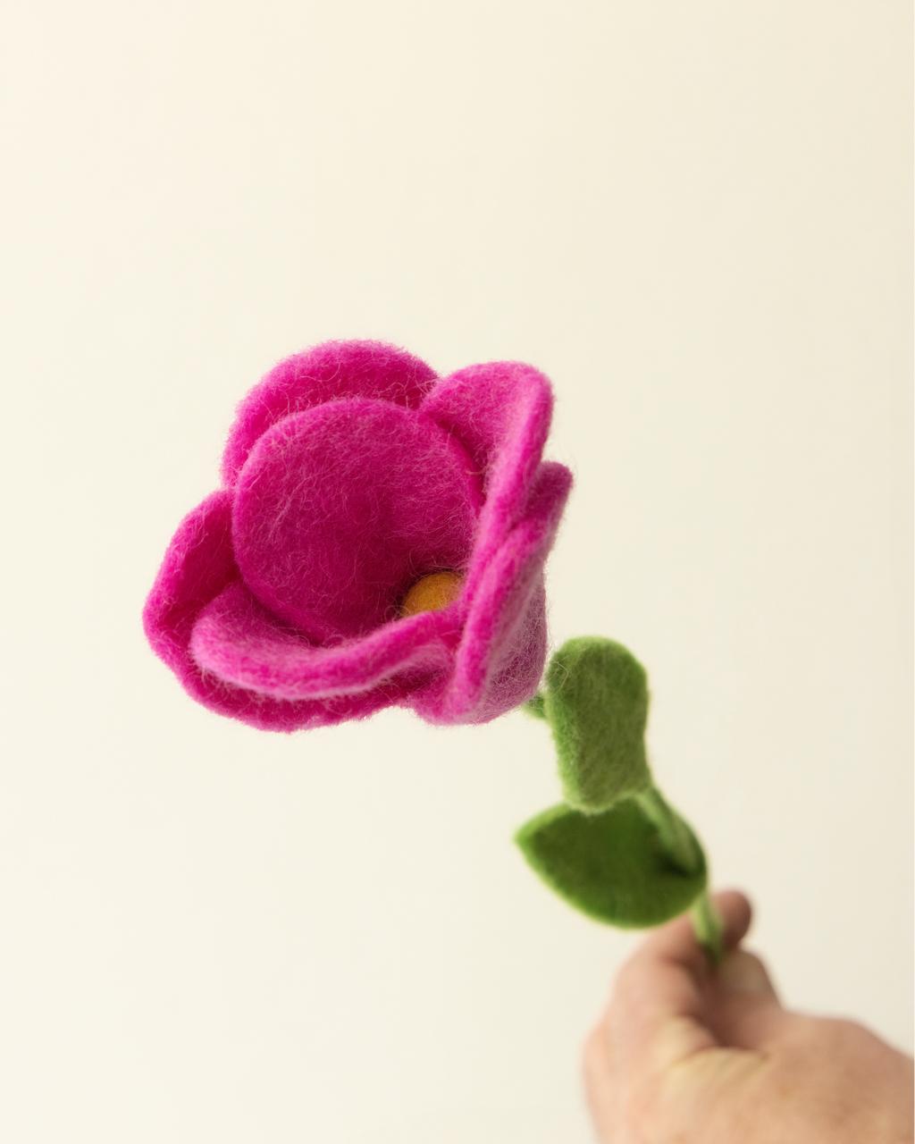 A hand holds a felted pink flower with green leaves and stem against a plain off-white background. 