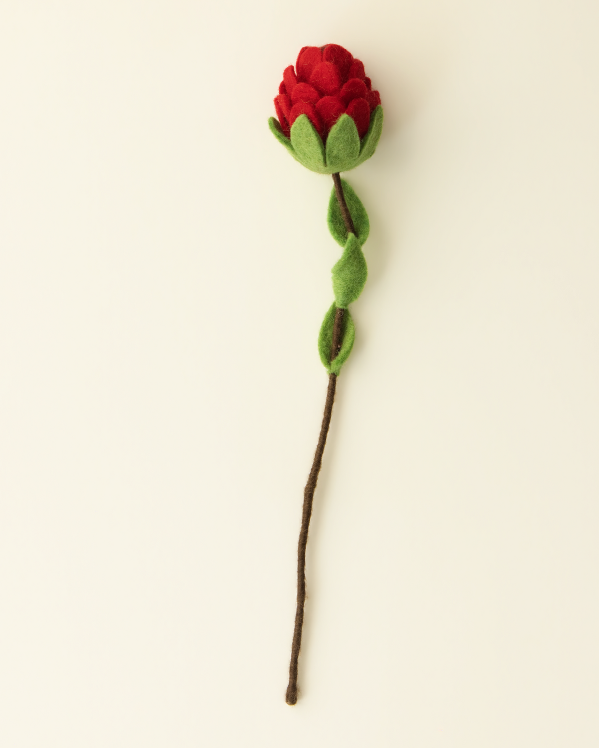 A single felt flower decoration with a red rosebud, green leaves, and a brown stem, photographed on a plain warm off-white background.