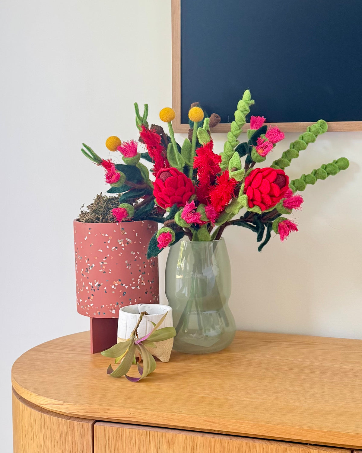 A vibrant artificial flower arrangement featuring bright red roses, pink blossoms, green cactus stems, and yellow pom-pom accents, displayed in a light green glass vase on a wooden console table next to a terracotta pot.